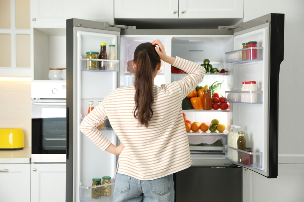 Back view of young woman standing in front of an open refrigerator in a kitchen.